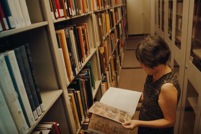 A woman holds an open book amid bookshelves in library stacks.